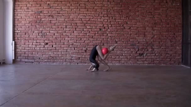 Rhythmische Sportgymnastik: Ein kleines Mädchen übt eine Gymnastikübung mit Ball. Zeitlupe. rote Ziegelwand auf dem Hintergrund — Stockvideo