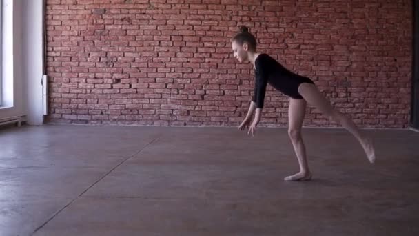 A flexible girl in a black bodysuit makes a gymnastic somersault in training studio with brick wall background, slow motion. Girl doing gymnastic flip. Acrobatic flip. Slow motion — Stock Video