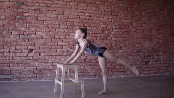 Красивая девочка-гимнастка в спортивном костюме делает разминку в студии и выполняет упражнения на растяжку мышц, опираясь на стул. Художественная гимнастика для девочек — стоковое видео