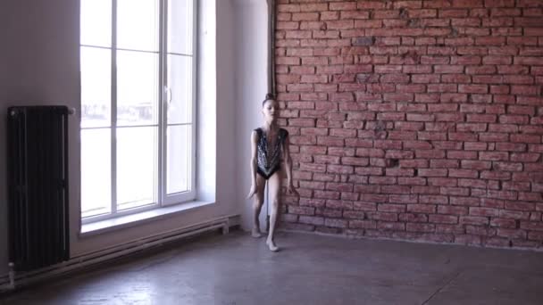 Linda chica dedicada a la gimnasia rítmica en traje de cuerpo artístico cerca de las ventanas. Joven gimnasta saltando en la escuela de gimnasia o estudio. Movimiento lento — Vídeo de stock