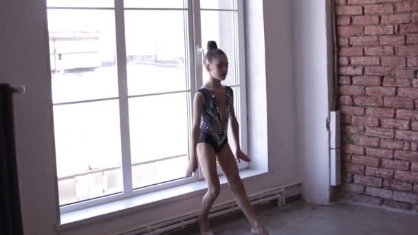 Chica joven artística dedicada a la gimnasia rítmica en traje de cuerpo artístico de pie cerca de las ventanas y preparándose para el salto. Entrenamiento de gimnasta joven en la escuela o estudio de gimnasia. En cámara lenta. Lado — Vídeo de stock