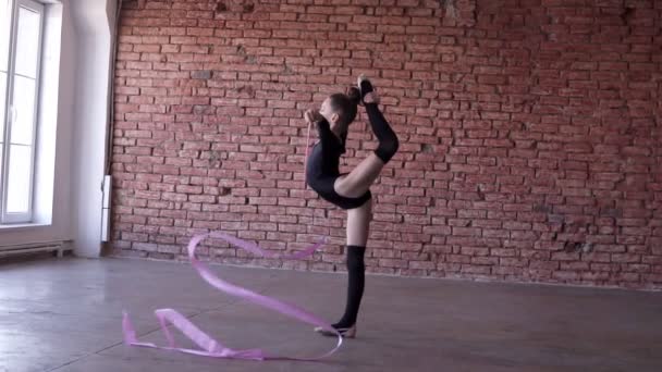 Gimnasta rítmica, niña en traje negro haciendo movimientos acrobáticos con la cinta - se coloca en una pose con la pierna extendida y agitando la cinta. Fondo de pared de ladrillo. Movimiento lento — Vídeos de Stock