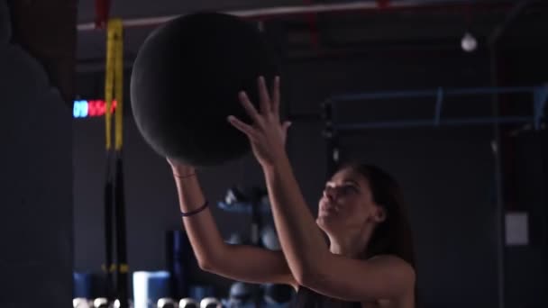 Activa, joven morena haciendo sentadillas mientras lanza una pelota de medicina contra una pared en un moderno gimnasio de color oscuro lleno de equipo. Vista de cerca — Vídeo de stock