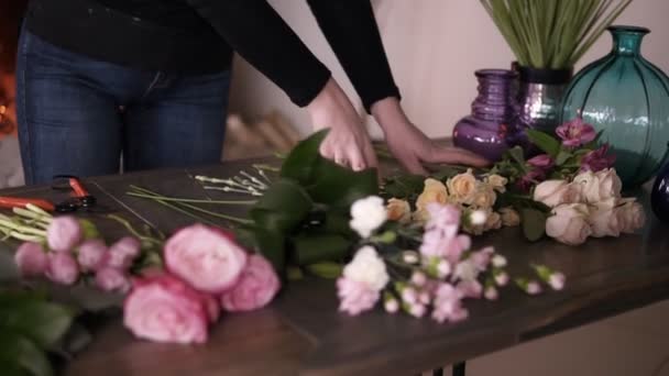 Longhaired μελαχρινή ανθοπωλείο προετοιμασία μελλοντικών μπουκέτο λουλούδια. Τακτοποίηση των διαφορετικών λουλουδιών στο τραπέζι - τουλίπες, τριαντάφυλλα, τριαντάφυλλα τσαγιού. Τζάκι στο φόντο — Αρχείο Βίντεο