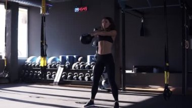 Spor fitness kız tam uzunlukta 10 kilo su ısıtıcısı çan ile crossfit egzersiz yapıyor. Başının altından büyük, siyah kettlebell'i kaldırıyor. Spor, arka planda spor ekipmanları