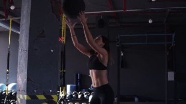 Siyah leeggings ve sütyen egzersiz genç bir fit kadın sideview görüntüleri arka planda ekipman dolu koyu renkli modern spor salonunda bir duvara bir ilaç topu atarak