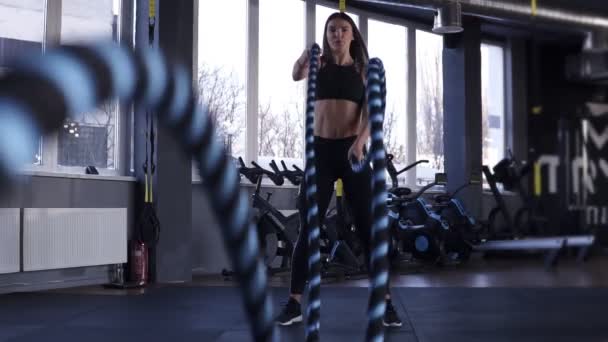Женщина тренируется в спортзале с боевыми канатами. Мощные привлекательные кавказские женщины делают боевые тренировки с веревками в тренажерном зале в замедленной съемке. Низкий угол съемки — стоковое видео