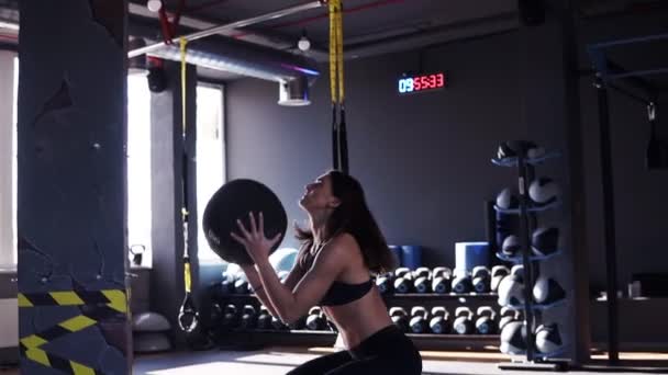 Filmato laterale di una giovane donna in forma che si allena lanciando una palla medica contro un muro in una moderna palestra di colore scuro piena di attrezzature sullo sfondo — Video Stock