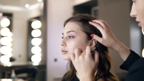 Mujer morena aplicando base o tóner en modelos cara y cuello con un gran pincel de maquillaje negro. Chica con un pelo largo marrón en el salón haciendo maquillaje, mirándose en el espejo. Maquillaje profesional — Vídeos de Stock