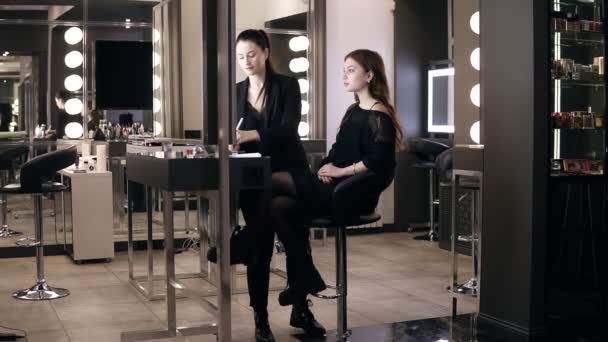 Hermosa joven en vestido negro casual sentado en la silla frente al espejo, mientras que el maestro de maquillaje femenino aplicando cosméticos en su cara usando un cepillo. Imágenes secundarias — Vídeos de Stock