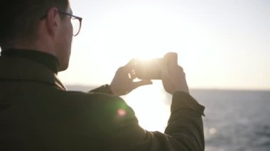 Sabah deniz sahilinde yürürken cep telefonu kullanarak şık kıyafetler giyen yetişkin adam, yükselen güneşin fotoğrafını çekiyor. Omzundan görüntüler. Güneş ışığı. Yavaş çekim