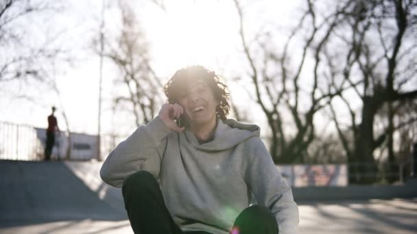 Joven alegre y sonriente está llamando a sus amigos usando el teléfono móvil mientras está sentado en una zona de skate en el parque de skate. Un tipo está sentado en su longboard en el suelo. El sol brilla en el fondo. Despacio. — Vídeo de stock