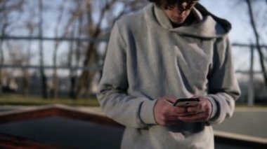 Young curly headed male skateboarder browsing in his mobile phone in the skate park on a sunny day. Lean on railing with his longboard next to him. The camera moves away from him for full length