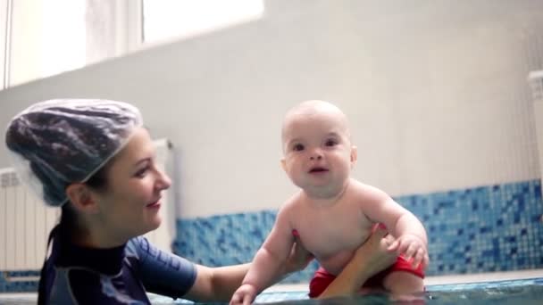 Un bambino carino in pantaloncini rossi si siede sul lato della piscina e poi si tuffa sott'acqua con l'aiuto di sua madre o allenatore. Una donna lo sostiene con le mani e tira in superficie — Video Stock