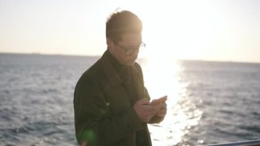 Gün batımında akıllı telefon mesajlaşma veya web sörf kullanarak kahverengi ceket ve şık gözlük çekici bir kafkas adam portresi. Telefona konsantre oldun. Deniz ve güzel sabah güneş ışığı üzerinde
