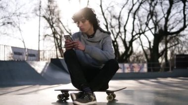 Genç adam kaykay parkında kaykay pistinde otururken cep telefonu tutuyor. Bir adam otururken akıllı telefonuyla internette sörf yapıyor. Arka planda güneş parlıyor