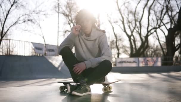 Mladý muž volá na přátele pomocí mobilního telefonu, zatímco sedí v bruslení zóny v skate parku. Chlápek sedí na dlouhém prkně na zemi. Slunce svítí na pozadí — Stock video