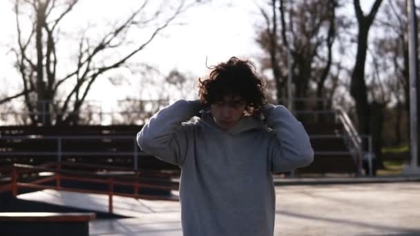 Portrait of a handsome young man with dark curly hair. Teen boy in grey hoodie walking over spring skate park with nature background. Guy walking confidently, putting hood up. Footage in slow motion — Stock Video