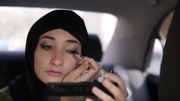 Retrato de una joven musulmana con pañuelo negro en la cabeza, sentada en el coche mientras mira un pequeño espejo cosmético y dibuja una línea con delineador de ojos. Es de día. Vista de cerca — Vídeo de stock