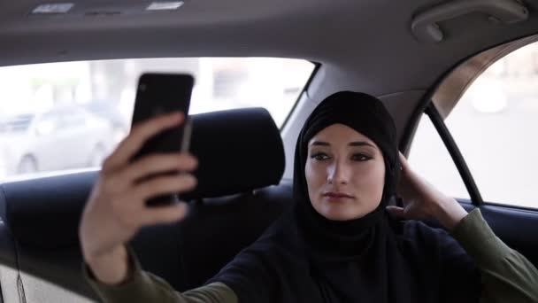 Портрет молодой, привлекательной мусульманки, носящей хиджаб в машине. Она одета в тёмную одежду, сидит на заднем сидении. Она использует свой мобильный телефон, делает селфи фото — стоковое видео