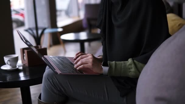 カフェで現代のラップトップに取り組んでいる集中若いイスラム教徒の女性。ヒジャーブの魅力的な女性は、彼女の膝の上にラップトップを保持し、インターネットで何かを探して、タイピング。側面図 — ストック動画