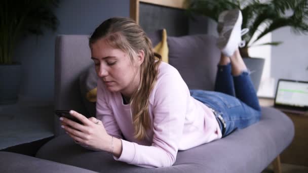 Menina atraente em suéter rosa usando telefone inteligente enquanto deitado no sofá na sala de estar. Digitar, assistir a vídeos ou navegar em mídias sociais no smartphone. Na frente do portátil dela. Movimento lento. Frente — Vídeo de Stock