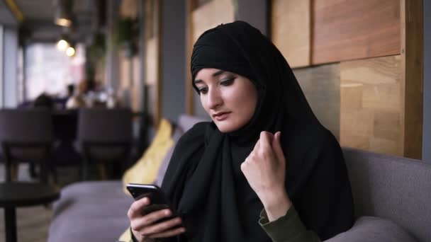 Donna musulmana in caffè utilizzando il suo smartphone, chattando navigazione online social media sharing lifestyle. Piacevoli, rilassanti messaggi di lettura. Donna vestita di hijab nero, velo. Lettura dal cellulare — Video Stock