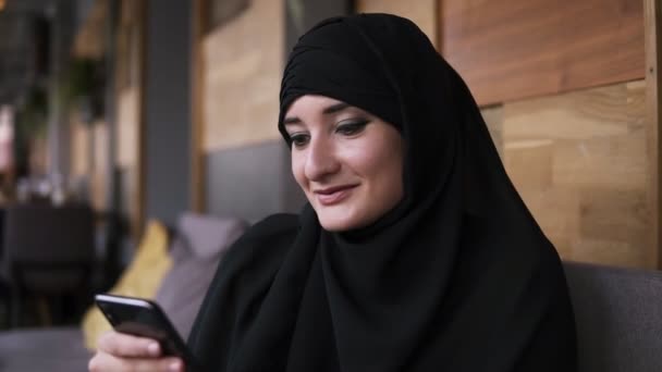 Κοντά στην μουσουλμανική γυναίκα στο καφέ χρησιμοποιώντας το smartphone της, συνομιλούν σε απευθείας σύνδεση με τους φίλους ή περιήγηση στα μέσα κοινωνικής δικτύωσης, μοιράζονται τον τρόπο ζωής. Απολαμβάνω, χαλαρώνω διαβάζοντας μηνύματα. Γυναίκα φορώντας μαύρη μαντίλα — Αρχείο Βίντεο