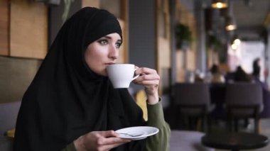 Modern kafede oturan siyah başörtüsü giyen genç müslüman kadın. Beyaz fincandan kahve içmek ve kafenin penceresinden sokağa düşünceli bir şekilde bakmak. Yakından kapatın. Yavaş çekim
