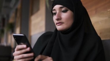 Arkadaş ya da sosyal medya tarama, yaşam tarzı paylaşımı ile online sohbet, onu Smartphone kullanarak kafede makyaj ile Müslüman kadın yakın. Zevk, rahatlatıcı mesajlar okuma. Siyah giyen kadın