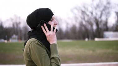Parkta bir akıllı telefonda arkadaşlarınızla sohbet geleneksel bir başörtüsü genç bir Müslüman kadın, bir Bayan bulutlu bir gün siyah hijab Park boyunca yürür