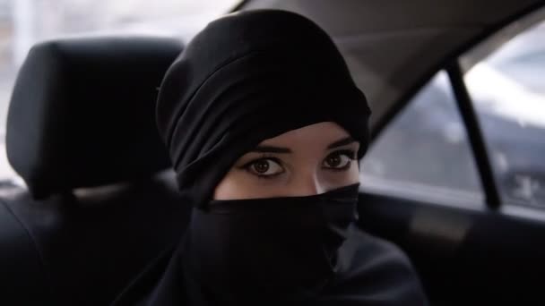 Portrait de jeune femme musulmane avec maquillage des yeux en niqab ou burqa noir, assise dans la voiture sur le siège arrière - regardant vers la caméra et souriant — Video