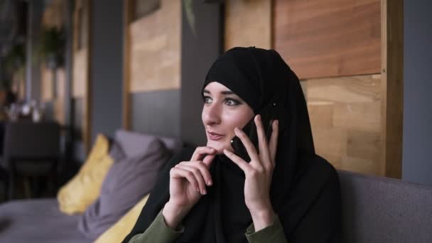 Joven mujer bastante musulmana con maquillaje en hijab hablando por teléfono y sonriendo, sentado en la cafetería, alegre. Vista lateral — Vídeo de stock