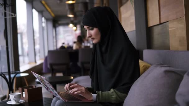 Konsentrat wanita muslim muda bekerja pada laptop modern di kafe. Wanita yang menarik mengenakan jilbab sambil memegang laptop sambil berlutut dan mengetik. Sisi tampilan. Gerakan lambat — Stok Video