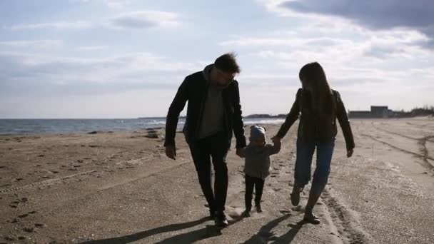 Современные, молодые родители с сыном посередине прогуливаются всю осень, весеннее побережье. Держась за руки, подбирайте детей, держа их за руки впереди. Счастливая семья. Вид спереди — стоковое видео