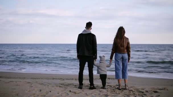 Bonita cena em câmera lenta de um jovem casal, com seu filho em pé em frente ao mar e olhando no horizonte. Vento, tempo frio — Vídeo de Stock