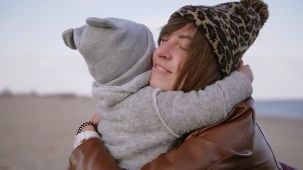 Mutlu sarılıyor ve kış leoparı güzel anne öpücüğü baskılı şapka ve deniz plajında açık havada onun yürümeye başlayan oğlu. Genç Anne Çocuk oğlunu kucaklıyor. Mutlu aile — Stok video