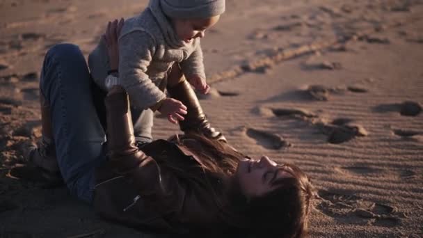 秋の服で幸せな、若い母親は砂の上に彼女の背中に横たわって、歌おうは彼女の子供を抱きしめ、手を伸ばし、笑顔で彼女を持ち上げます。日当たりの良い、幸せな一日屋外 — ストック動画