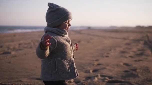 秋のビーチで抱き合って母親に走っている幸せな小さな幼児。かわいい子供は彼のお母さんを抱きしめ、彼らは砂の上に落ちます。屋外で子供、幸せな家族、幸せな子供の頃のコンセプトママ — ストック動画