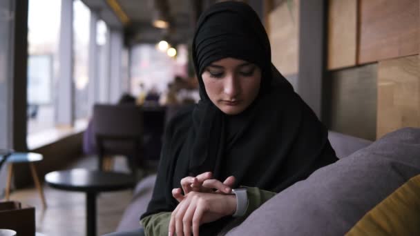 Retrato de mulher muçulmana em hijab preto sentado no café moderno sozinho e usando seus relógios inteligentes, deslize os dados com o dedo. Movimento lento. Fundo desfocado — Vídeo de Stock