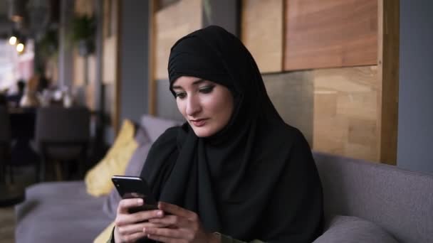 Mujer musulmana en la cafetería usando su teléfono inteligente, charlando en línea navegando por las redes sociales compartiendo estilo de vida. Disfrutando, relajando los mensajes de lectura. Mujer con hijab negro, pañuelo en la cabeza — Vídeos de Stock