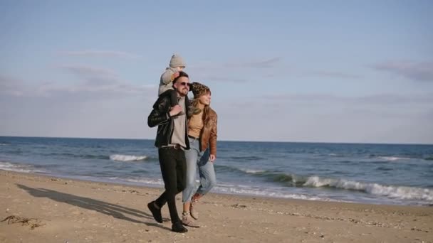 Молодой отец носит сына в плече, молодая семья гуляет по берегу моря, наслаждается совместными выходными на природе. Влюблённая женщина смотрит на мужчину. Любовь, семья, концепция отдыха — стоковое видео