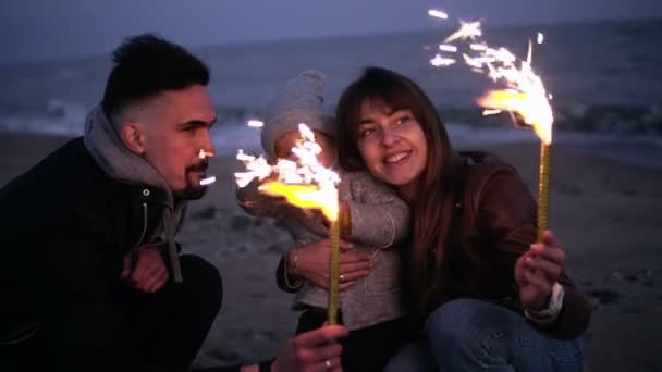 Bambina sulle braccia dei padri che guarda sua madre, il padre che agita scintille sulla spiaggia al tramonto della sera. Famiglia giovane e felice, viso felice, mare. Rallentatore — Video Stock