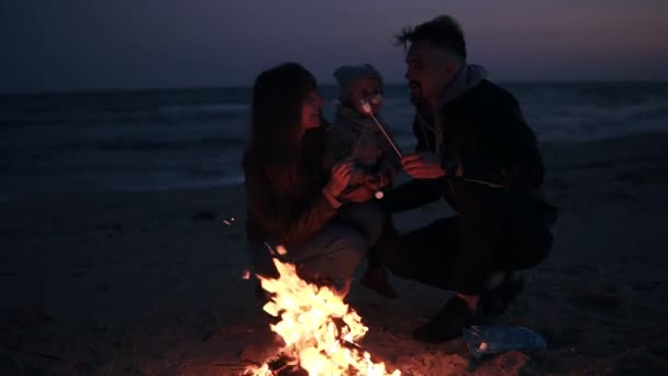 Mamma, papà in abiti caldi stanno arrostendo marshmallow su bastoncini di legno con il loro bambino. Falò sulla spiaggia la sera. Si nutrono a vicenda di bastone, prendendo in giro. Rallentatore — Video Stock