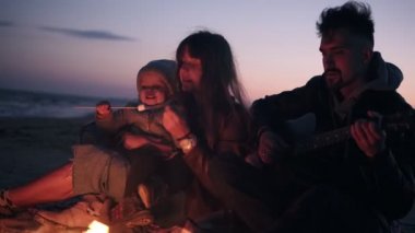 Şenlik ateşinin yanında oturan üç kişilik sevimli, beyaz aile - gitar çalan baba, sevimli bebeğini kavrulmuş marshmallow ile besleyen ve ellerini ateşle ısıtan genç uzun saçlı kadın