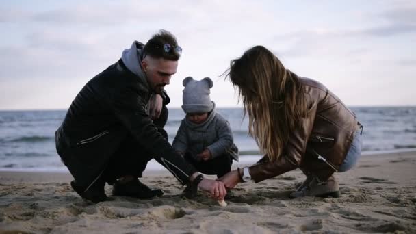 Mãe e pai com seu filho menino criança brincando com a areia em tempo vento nublado frio, o homem ajuda pequeno menino cavando buraco na areia. Vista frontal — Vídeo de Stock