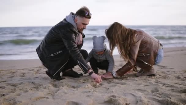 Les jeunes parents avec leur enfant tout-petit garçon jouant avec le sable par temps froid nuageux et venteux, l'homme et la femme aident le petit garçon à creuser un trou dans le sable, en dessinant des lignes sur le sable. Vue de face — Video