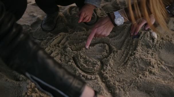 Крупный план съемки молодых родителей со своим ребенком малышом, играющим с песком в холодную облачную погоду, мужчина и женщина помогают своему сыну рисовать сердце на песке, украшая его морскими раковинами — стоковое видео