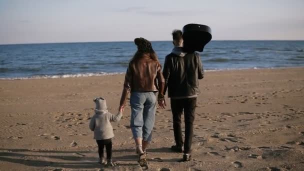 Paseos familiares por la playa. Los padres caminan con su pequeña hija o el sol a lo largo de la orilla del mar. Se toman de la mano. Están contentos. Padre sosteniendo una guitarra en la mano. En cámara lenta. Vista rara — Vídeo de stock