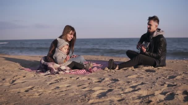 Retrato de uma família senta-se em um cobertor xadrez na praia como o pai toca guitarra e mãe com a criança ouvindo-o. Tempo frio, seasisde. Movimento lento — Vídeo de Stock